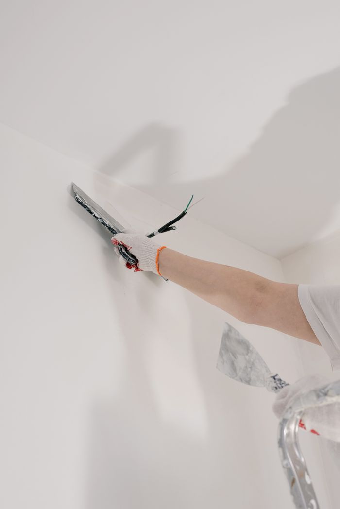 Crop unrecognizable male in gloves holding tool while standing on ladder and renovating walls in flat